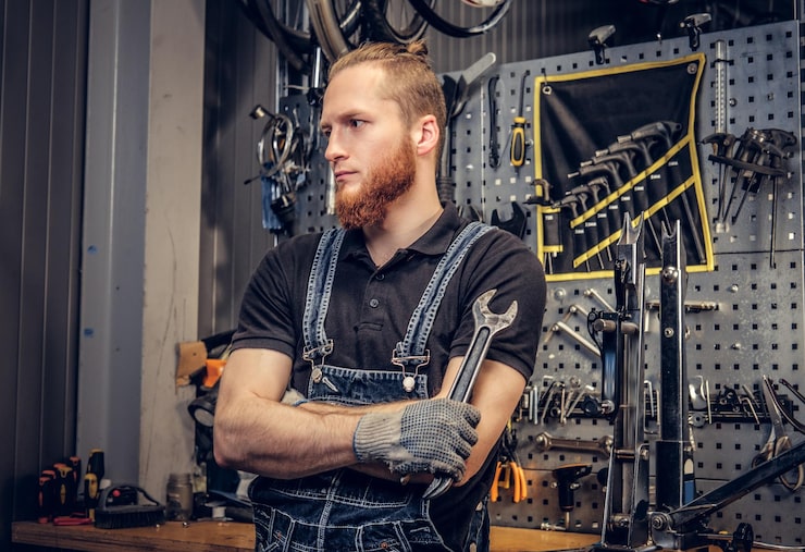 Bezpłatny plik Zdjęcie portret brodaty mechanik rowerowy ze skrzyżowanymi rękami trzyma klucz do kubka na tle stoiska narzędziowego w warsztacie.