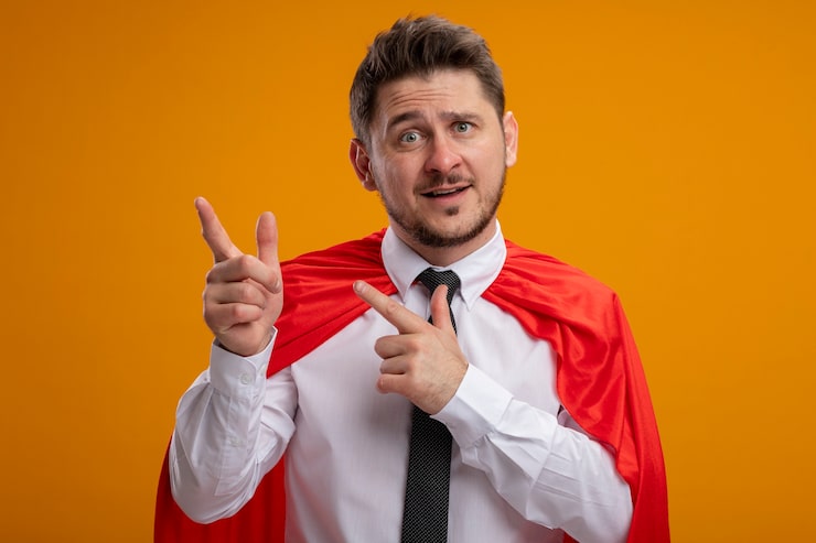 Bezpłatny plik Zdjęcie superbohater biznesmen w czerwonej pelerynie, wskazując palcami wskazującymi na bok uśmiechnięty stojący na pomarańczowym tle