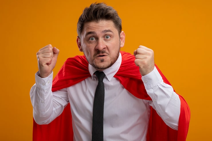 Bezpłatny plik Zdjęcie superbohater biznesmen w czerwonej pelerynie zaciskając pięści z gniewną twarzą stojącą na pomarańczowej ścianie