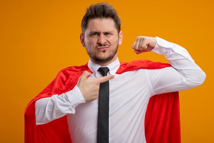 Bezpłatny plik Zdjęcie zadowolony biznesmen superbohatera w czerwonej pelerynie podnoszący pięść wskazujący palcem wskazującym na bicepsy pokazujący siłę wyglądającą pewnie stojąc na pomarańczowym tle