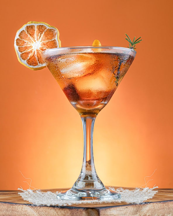 Foto gratuito cóctel de naranja en copa de martini con ramita de romero y limón deshidratado sobre superficie de madera rústica