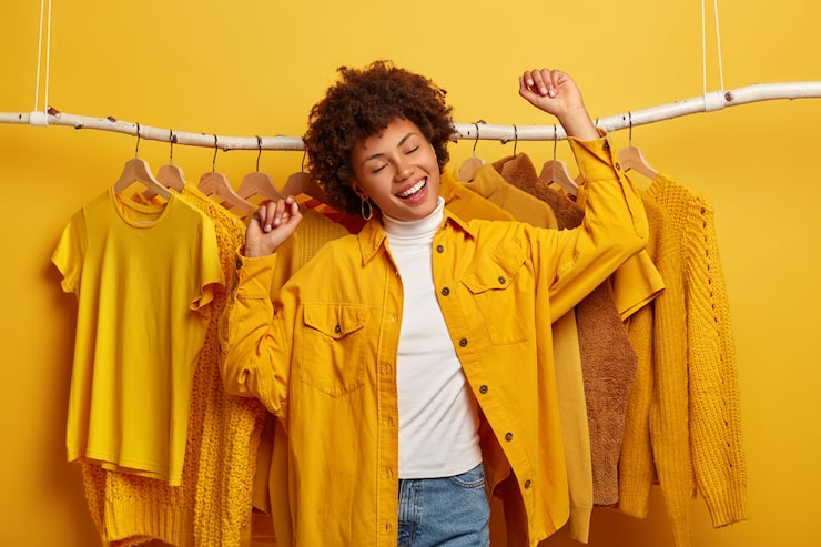 Foto gratuito compradora de ropa de mujer rizada despreocupada baila con felicidad, levanta los brazos, compra ropa amarilla de la nueva colección, se regocija con el día de compras exitoso, está en alto espíritu, baila contra los atuendos en el estante