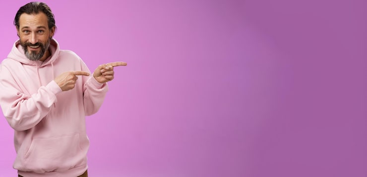 Foto gratuito descarado, confiado y apuesto hombre macho con capucha rosa sonriendo ampliamente invitando a unirse apuntando a la derecha