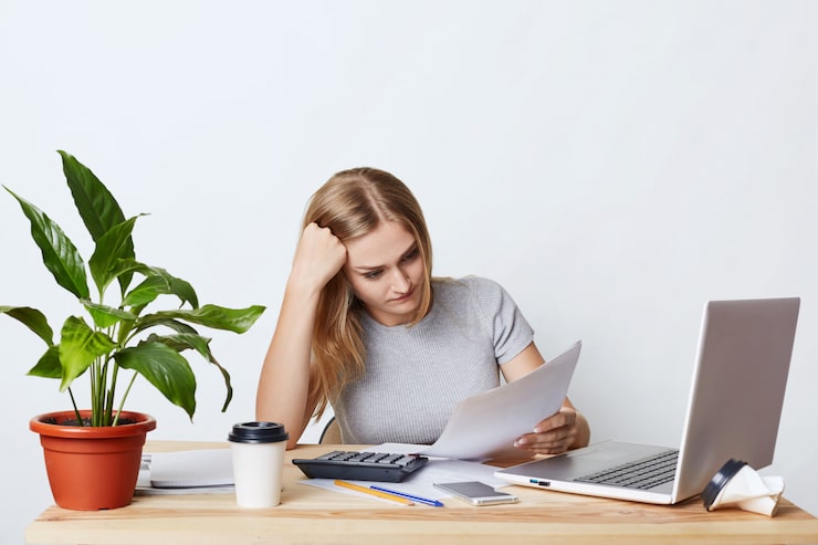 Foto gratuito empresaria con exceso de trabajo sentada en una mesa de madera, rodeada de aparatos modernos, leyendo documentos atentamente, tratando de entender todo. mujer contadora calculando facturas y gastos