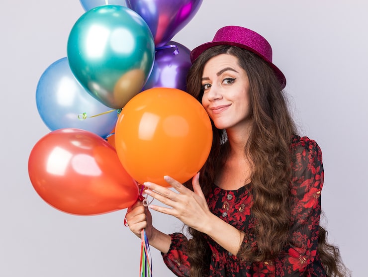 Foto gratuito feliz joven fiestera con sombrero de fiesta sosteniendo y tocando globos mirando a la cámara aislada en la pared blanca