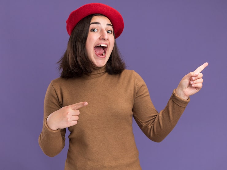 Photo Gratis gadis kaukasia muda cantik yang bersemangat dengan topi baret menunjuk ke samping di atas latar belakang ungu