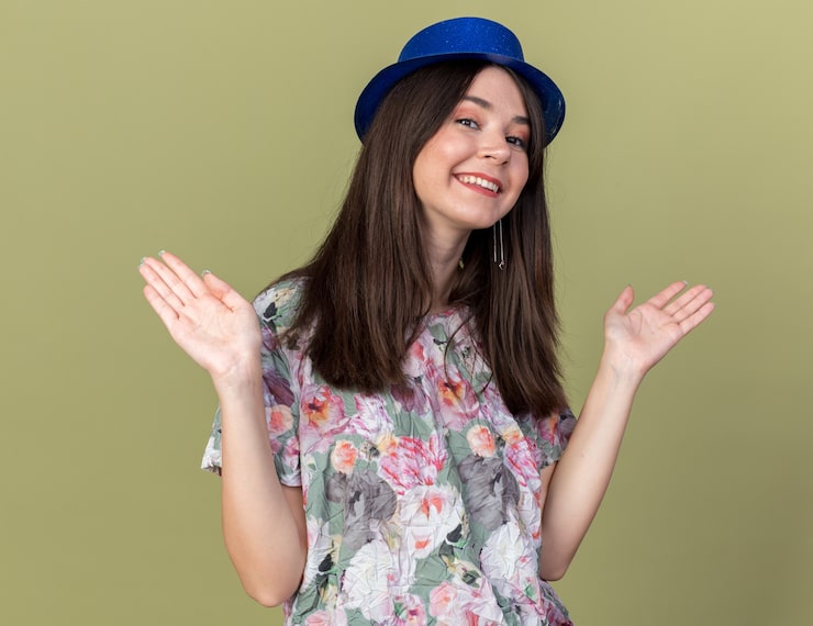 Photo Gratis gadis muda cantik tersenyum mengenakan topi pesta merentangkan tangan yang terisolasi di dinding hijau zaitun