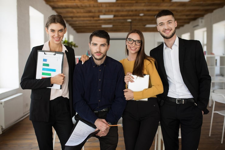 Foto gratuito grupo de jóvenes empresarios sonrientes con trajes clásicos mirando alegremente a la cámara juntos mientras pasan tiempo en el trabajo en una oficina moderna