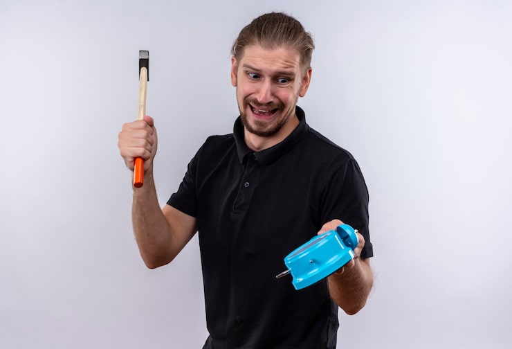 Foto gratuito hombre guapo joven decepcionado en camisa polo negra sosteniendo el reloj de alarma y balanceando un martillo que va a romper el reloj de pie sobre fondo blanco.