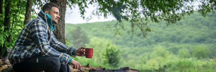 Foto gratuito un hombre con una taza de bebida caliente se calienta junto al fuego en el bosque