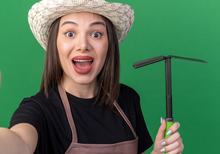 Foto gratuito jardinero femenino bastante caucásico sorprendido que lleva el sombrero que cultiva un huerto que sostiene el rastrillo de la azada que mira la cámara en verde
