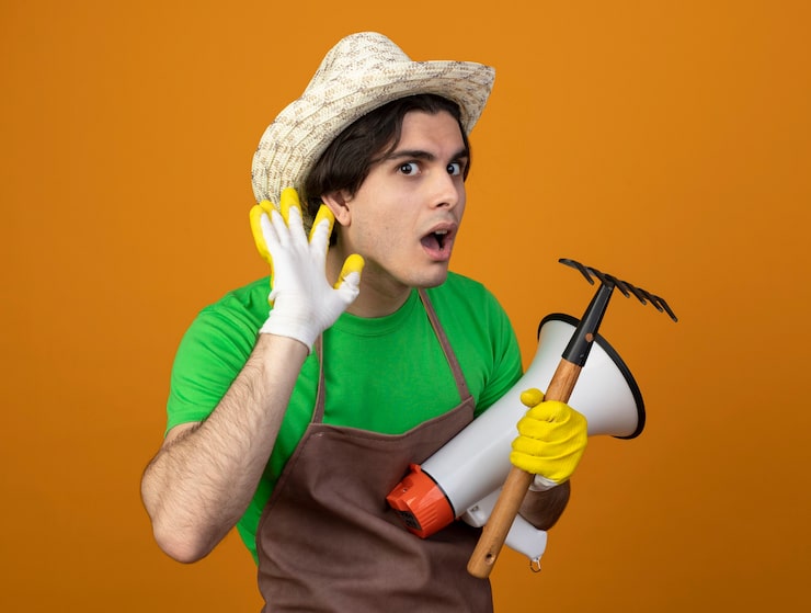 Foto gratuito jardinero de sexo masculino joven sospechoso en uniforme con sombrero de jardinería con guantes sosteniendo altavoz con rastrillo mostrando gesto de escuchar aislado en naranja