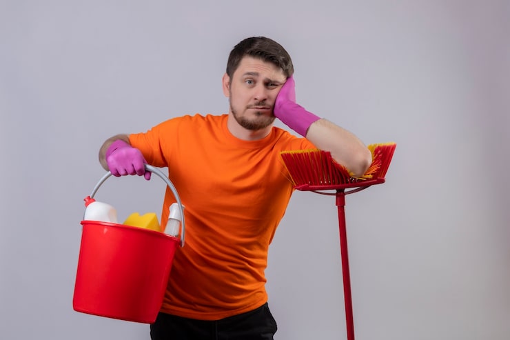 Foto gratuito joven vestido con camiseta naranja y guantes de goma sosteniendo un balde con herramientas de limpieza y un trapeador con aspecto cansado y aburrido de pie sobre la pared blanca
