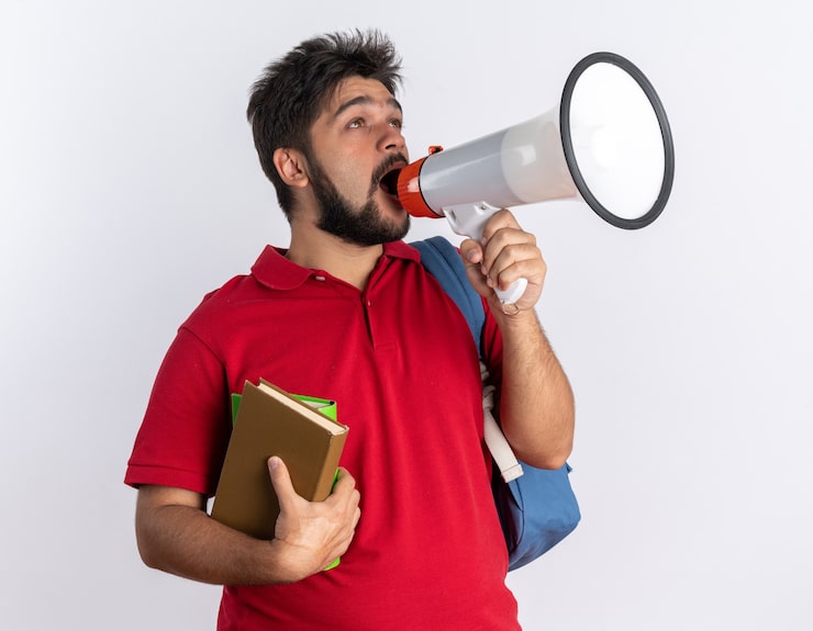 Photo Gratis mahasiswa muda berjenggot emosional dengan kemeja polo merah dan tas ransel memegang buku catatan berteriak ke megafon berdiri di atas dinding putih