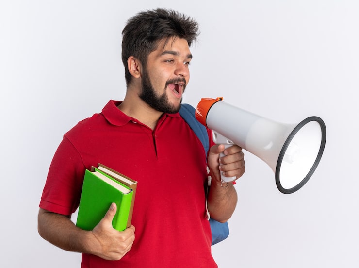 Photo Gratis mahasiswa muda berjenggot mengenakan kaos polo merah dengan ransel memegang buku catatan berteriak ke megafon bahagia dan positif berdiri