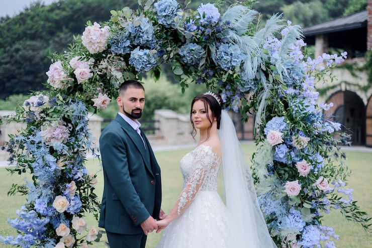 Photo Gratis pasangan pengantin kaukasia yang cantik berdiri di depan gerbang lengkung yang dihiasi dengan hydrangea biru dan berpegangan tangan bersama