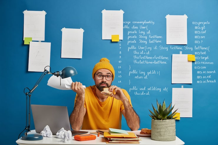 Photo Gratis pekerja lepas berjanggut yang bingung mengembangkan proyek startup, makan siang, makan camilan lezat, memegang dokumen kertas di tangan, berpose di desktop.