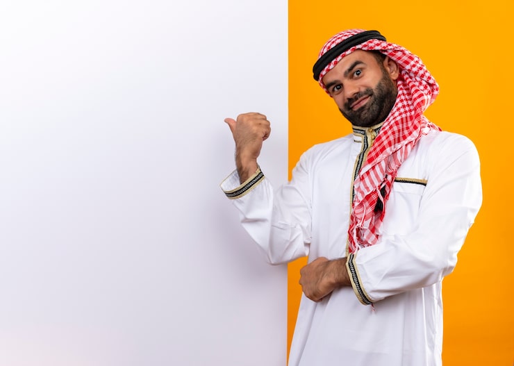 Photo Gratis pengusaha arab dengan pakaian tradisional berdiri di dekat papan iklan kosong menunjuk dengan jari ke arahnya dengan senyum percaya diri di wajahnya di depan dinding oranye