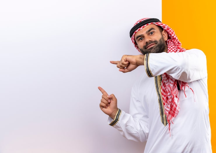 Photo Gratis pengusaha arab mengenakan pakaian tradisional berdiri di dekat papan reklame kosong menunjuk dengan jari ke sana dengan senyum percaya diri di wajah di atas dinding oranye