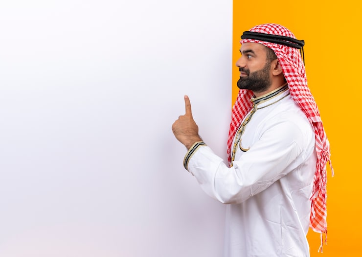 Photo Gratis pengusaha arab mengenakan pakaian tradisional dengan papan reklame kosong putih menunjuk jari ke atas tersenyum berdiri di depan dinding oranye