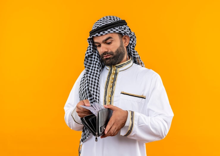 Photo Gratis pengusaha arab mengenakan pakaian tradisional melihat uang tunai di dompetnya dengan wajah serius berdiri di depan dinding oranye