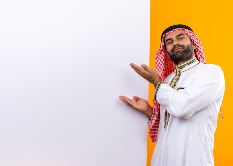 Photo Gratis pengusaha arab mengenakan pakaian tradisional mempersembahkan dengan lengan papan reklame kosong berdiri di depan dinding oranye