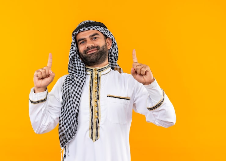 Photo Gratis pengusaha arab mengenakan pakaian tradisional menunjuk dengan jari telunjuk ke atas tersenyum percaya diri berdiri di depan dinding oranye