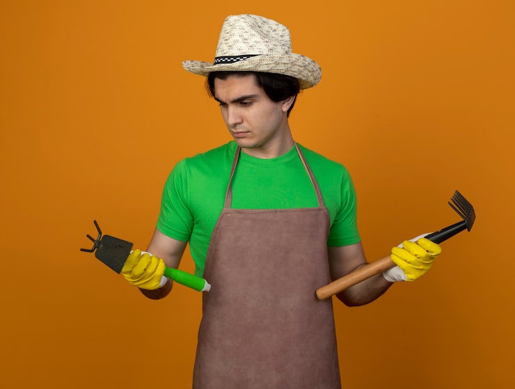 Foto gratuito pensando joven jardinero en uniforme con sombrero de jardinería con guantes sosteniendo y mirando azada rastrillo con rastrillo aislado en la pared naranja