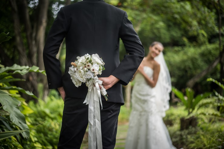 Photo Gratis potret seorang pengantin pria menyembunyikan buket bunga di belakang punggungnya untuk mengejutkan pengantin wanita.