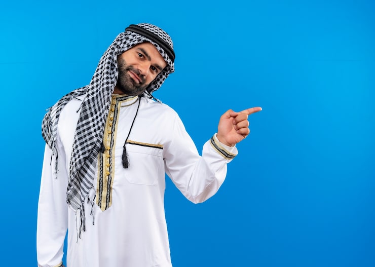 Photo Gratis pria arab mengenakan pakaian tradisional dengan senyum di wajah menunjuk dengan jari ke samping berdiri di depan dinding biru