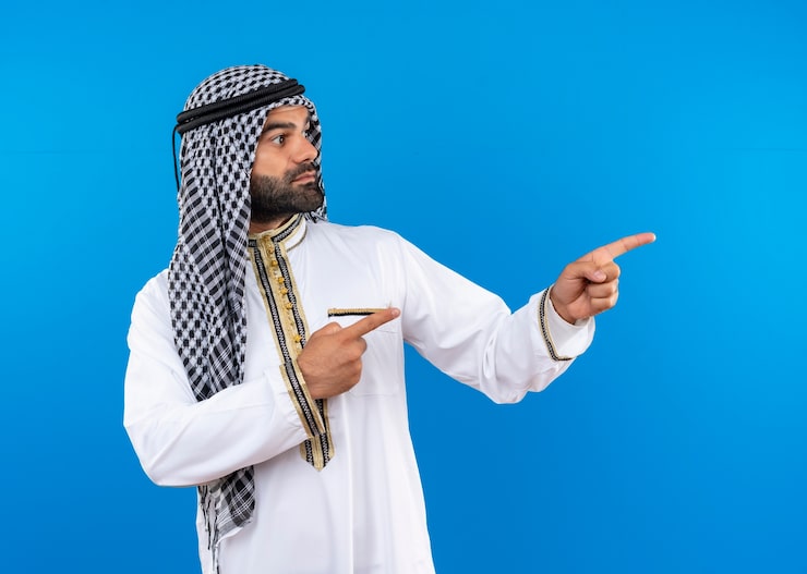 Photo Gratis pria arab mengenakan pakaian tradisional melihat ke samping menunjuk dengan jari ke samping dengan wajah serius berdiri di depan dinding biru