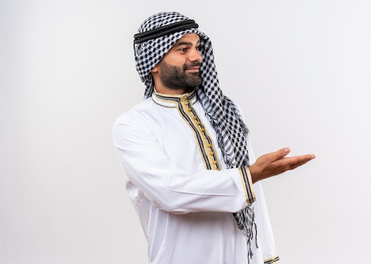 Photo Gratis pria arab yang senang dengan pakaian tradisional melihat ke samping mempersembahkan ruang salinan dengan lengan tangan berdiri di atas dinding putih