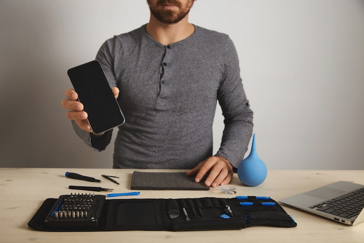 Photo Gratis profesional berjanggut menunjukkan smartphone yang diperbaiki setelah penggantian layanan, di atas alat spesifiknya di tas peralatan di dekat laptop di atas meja putih kayu