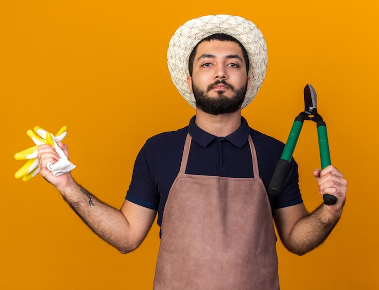 Foto gratuito seguro joven jardinero varón caucásico vistiendo sombrero de jardinería sosteniendo guantes y tijeras de jardinería aislado en la pared naranja con espacio de copia