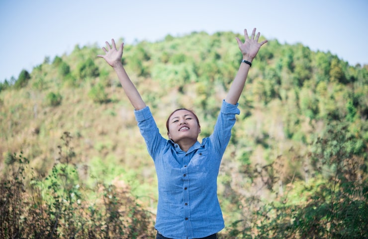 Photo Gratis seorang wanita muda berdiri menghadap langit dengan tangan terangkat saat mendaki gunung.