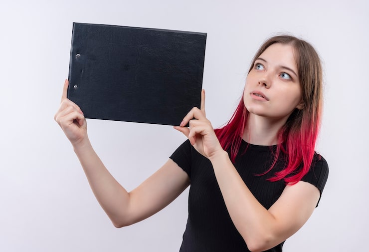Photo Gratis wanita muda cantik terkesan mengenakan t-shirt hitam memegang clipboard di dinding putih yang terisolasi