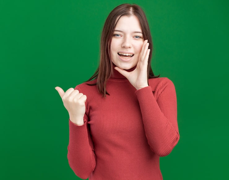 Photo Gratis wanita muda cantik yang riang menunjuk ke samping sambil memegangi tangan di dekat mulut berbisik