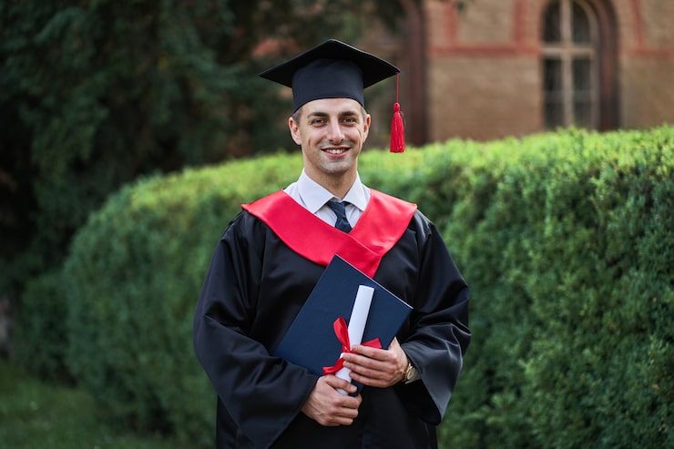 Foto gratuito felice maschio caucasico laureato in bagliore di laurea con diploma che guarda l'obbiettivo nel campus.