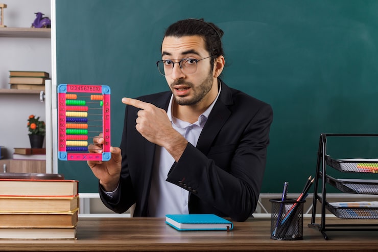Foto gratuito insegnante maschio sorpreso con gli occhiali che tengono e indica l'abaco seduto al tavolo con gli strumenti della scuola in classe