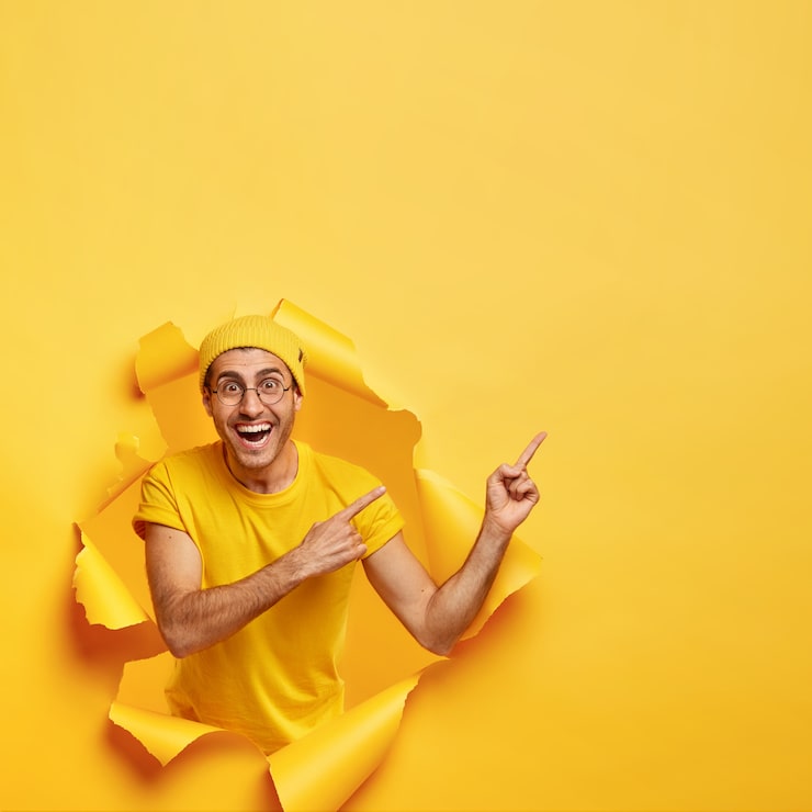Foto gratuito l'immagine luminosa dell'uomo felice ha un sorriso a trentadue denti, punta sullo spazio libero, pone in un muro di carta strappata