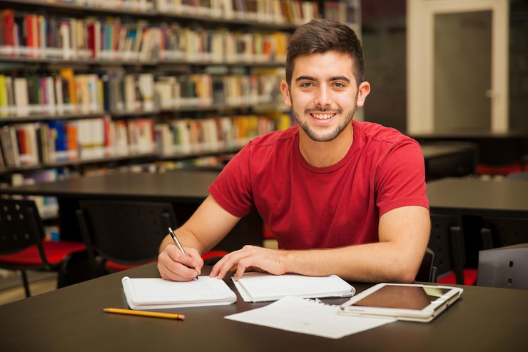 Foto gratuito studente universitario maschio attraente che fa dei compiti nella biblioteca della scuola e che sorride