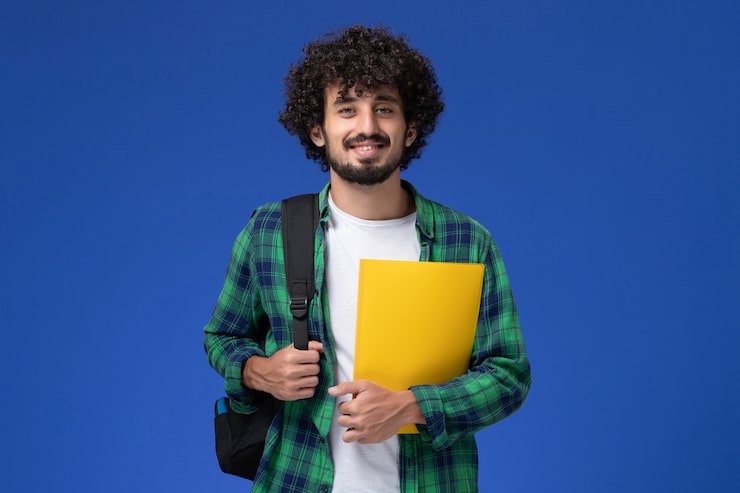 Foto gratuito vista frontale dell'allievo maschio in camicia a scacchi verde che porta zaino nero e che tiene i file sulla parete blu