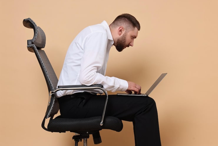 Photo pria dengan postur tubuh buruk duduk di kursi dan menggunakan laptop dengan latar belakang oranye pucat