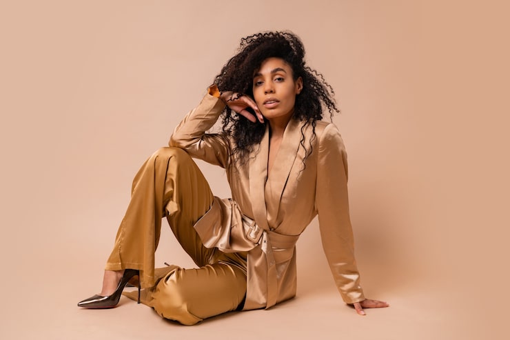 Foto grátis linda mulher negra com lindos cabelos ondulados em um elegante terno de cetim dourado posando sobre uma parede bege. look da moda primavera.