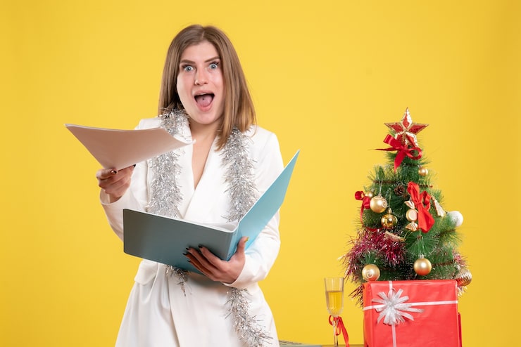 Foto grátis médica de frente, de pé, segurando documentos na mesa amarela com uma árvore de natal e caixas de presente