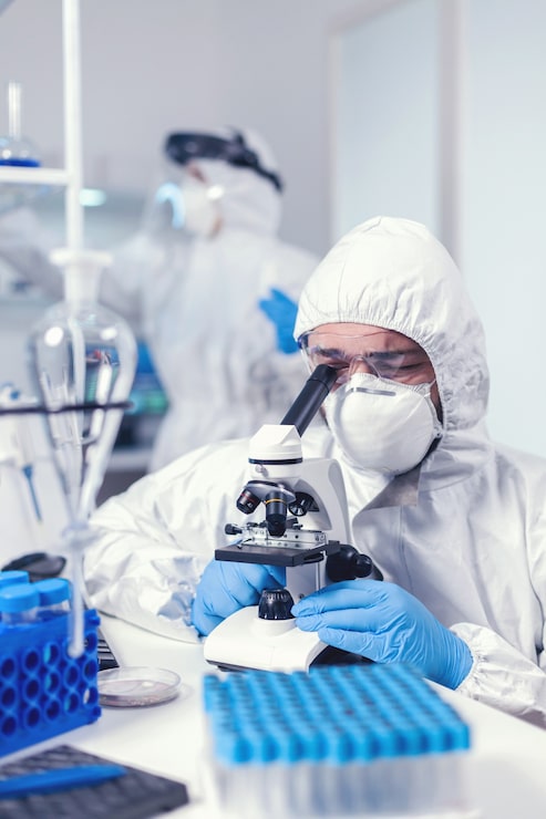 Foto grátis químico com máscara facial e vacina de desenvolvimento de ppe para cobiçado olhando através do microscópio. cientista em traje de proteção, sentado no local de trabalho, usando tecnologia médica moderna durante a epidemia global.