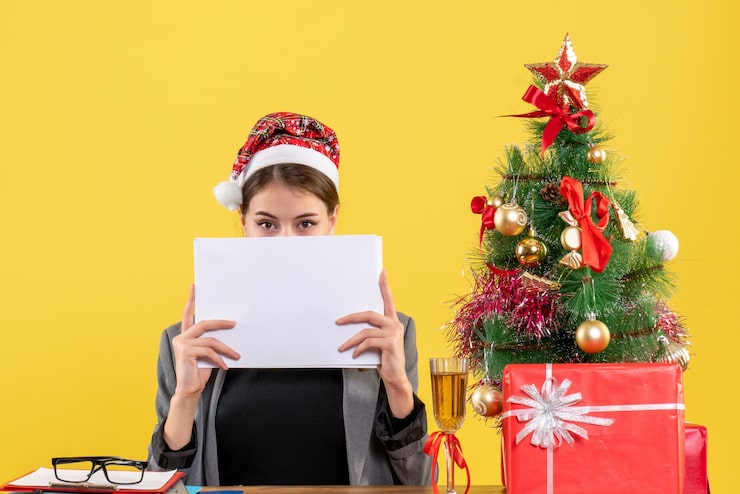 Foto grátis vista frontal jovem com chapéu de natal sentada à mesa, cobrindo o rosto com documentos árvore de natal e coquetel de presentes