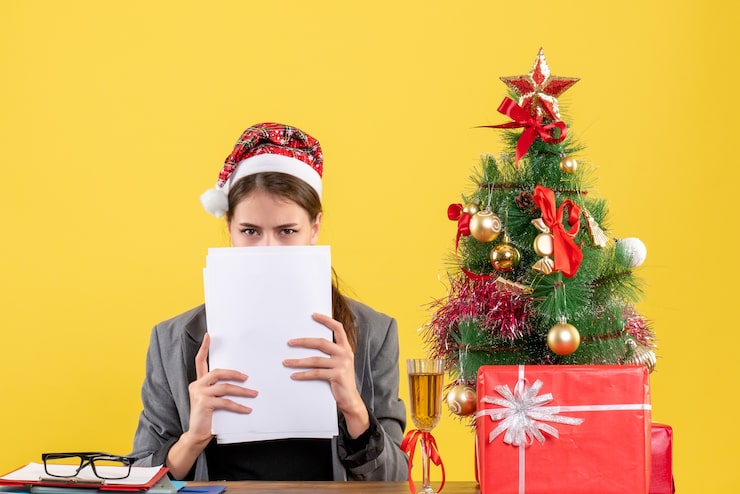 Foto grátis vista frontal jovem com chapéu de natal sentada à mesa, cobrindo o rosto com documentos árvore de natal e coquetel de presentes