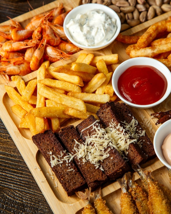 Foto grátis vista superior de petiscos variados de cerveja como palitos de pão frito com pistache de batatas fritas de queijo e camarão cozido com molhos em uma placa de madeira