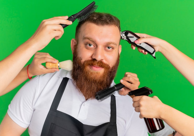 Kostenlose Foto bärtiger friseur in der schürze lächelnd, seinen haarschnitt und seinen bart geformt von anderen händen über grüner wand